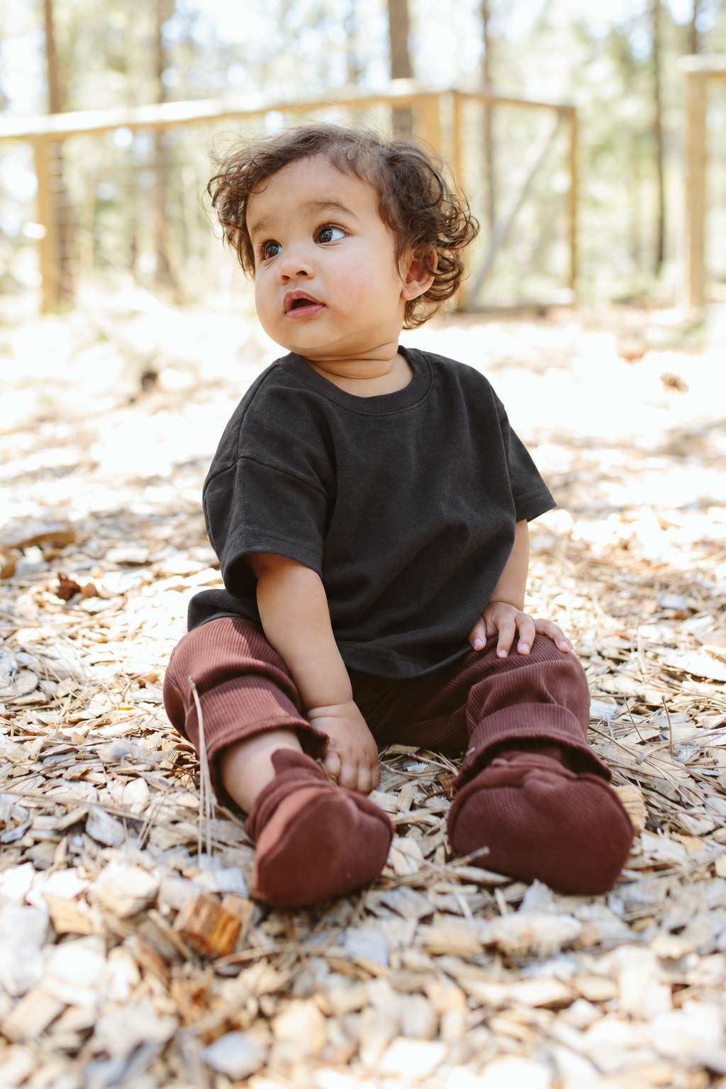 OVERSIZED TEE in asphalt black worn by a toddler sitting outdoors on wood chips