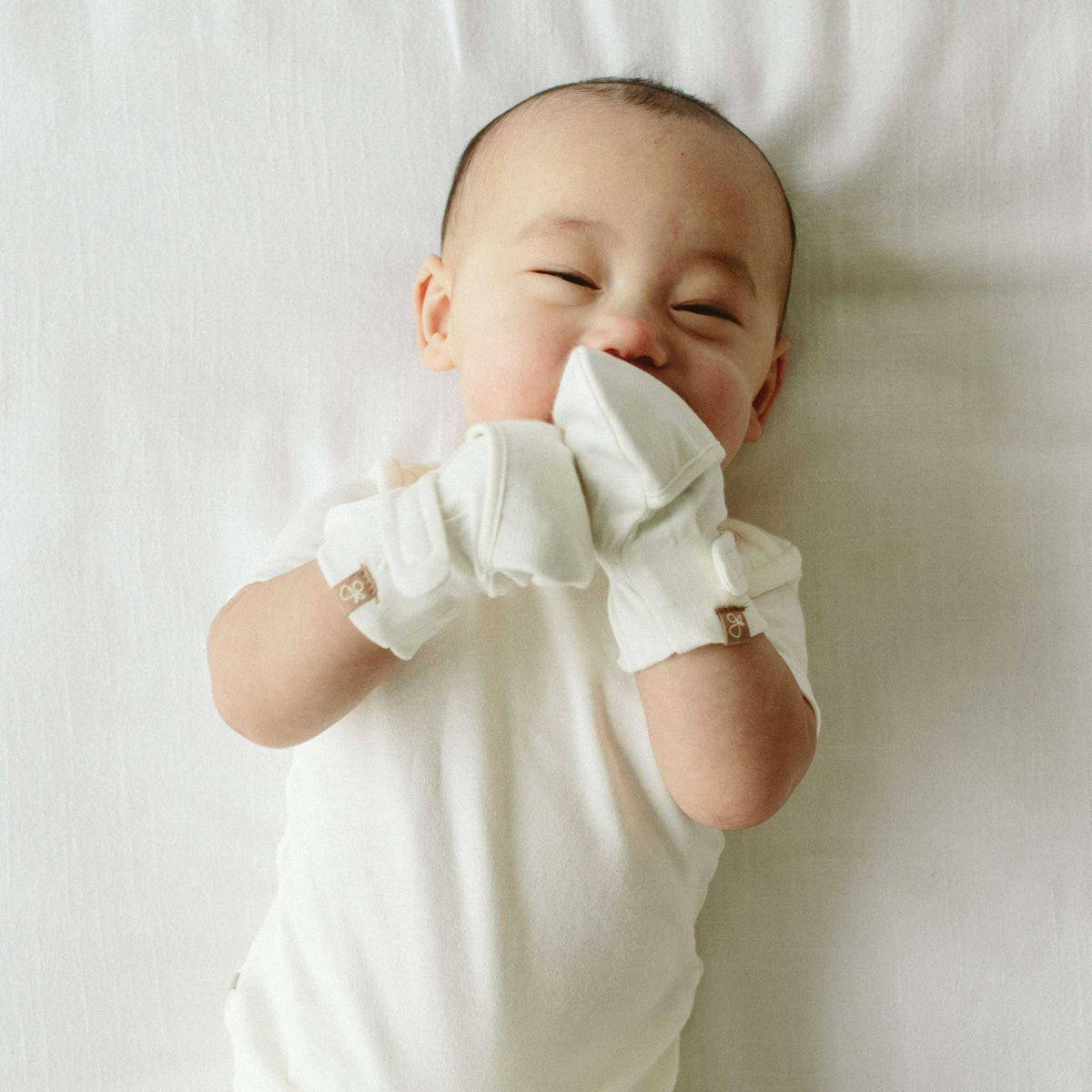 STAY ON MITTS are soft white baby mittens with adjustable wrist closures, worn by an infant covering their hands