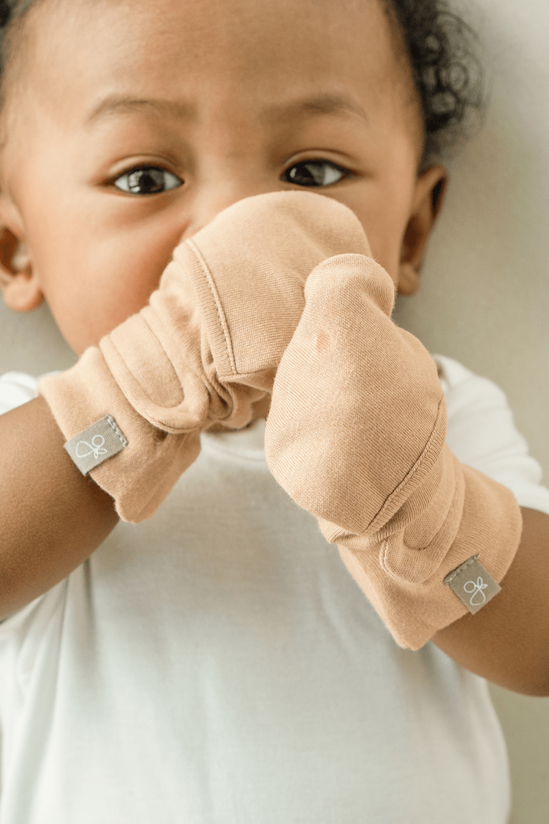 STAY ON MITTS in soft sand-colored fabric, worn by a baby with adjustable wraparound cuffs and small logo tabs